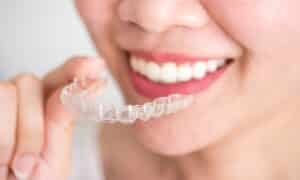 Close-up of a smiling woman holding a clear Invisalign aligner. The image showcases the aligner and her straight, white teeth. The setting is bright and focused on her mouth and hand.
