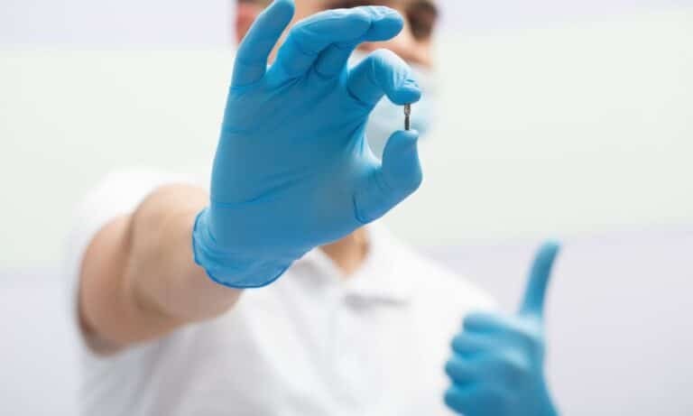 Dentist holding a small dental implant with a gloved hand, giving a thumbs-up gesture with the other hand. The background is blurred, focusing on the implant and the positive gesture.