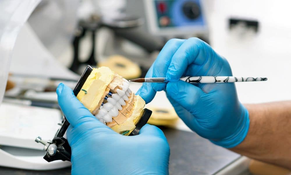 A dental technician wearing blue gloves works on a dental crown model with a brush in a lab setting, showcasing the detailed process of creating dental crowns. The image illustrates the precision and craftsmanship involved in dental crown fabrication.