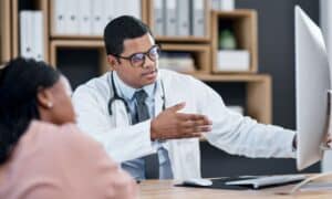 Doctor explaining dental treatment options to a patient using a computer screen. The doctor is wearing a white coat, glasses, and a stethoscope, while the patient listens attentively. The background shows shelves with binders and books.