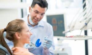 Dentist explaining the Invisalign® treatment to a smiling young patient. The dentist is holding a clear aligner and a dental tool, wearing gloves and protective eyewear. The patient is sitting in a dental chair, and there is a computer screen and dental equipment in the background.