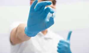 A dentist wearing blue gloves holding a small dental implant screw, giving a thumbs-up gesture in the background, illustrating the concept of dental implants.