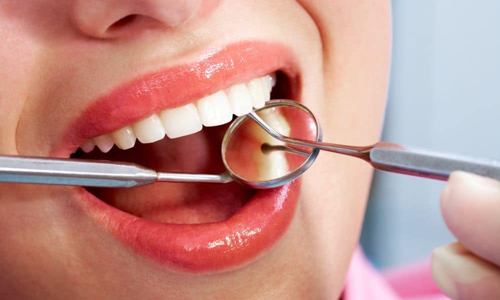 Close-up of a person's mouth during a dental examination. The dentist is using a dental mirror and a probe to check the teeth. The patient has healthy, clean teeth and is wearing lipstick.