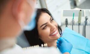 Dentist showing a patient a dental crown sample to match the color of her teeth. The patient is smiling and sitting in a dental chair, while the dentist, wearing gloves and a mask, holds the sample. The background is a dental clinic with equipment visible.