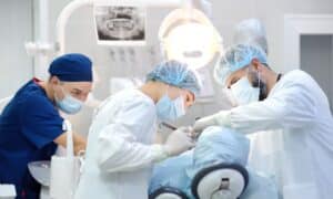 Dental surgeons performing an implant procedure on a patient in a dental clinic. The surgeons are wearing masks, gloves, surgical caps, and gowns, working under a bright overhead light. An X-ray image is visible in the background.