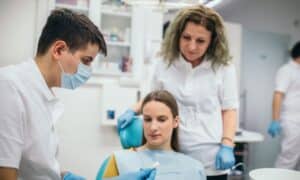 Dentist explaining the dental implant procedure to a patient, with another dental professional observing. The patient is seated in a dental chair, wearing a bib, while the dentist holds a model of the implant. All individuals are wearing masks and gloves. The background is a dental clinic.