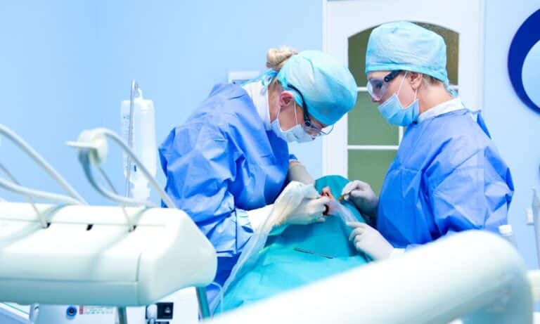 Dental surgeons performing an All-on-4 implant procedure on a patient. Both surgeons are wearing blue surgical gowns, masks, and gloves, working under a bright overhead light in a sterile clinical environment. Dental equipment is visible in the background.