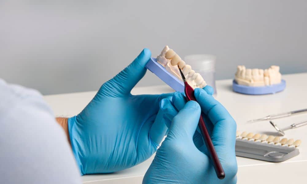 Dental technician working on a dental crown model, using a tool to shape and refine it. The technician is wearing blue gloves, and the workspace includes additional dental models, tools, and a shade guide. The background is a clean, clinical setting.
