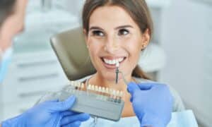 Dentist comparing tooth shade samples with a patient's teeth during a consultation. The patient is smiling, and the dentist is holding a shade guide and a dental tool, both wearing gloves. The setting is a dental clinic.