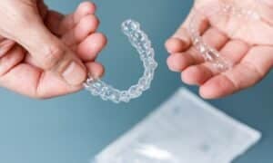 Two hands holding clear Invisalign® aligners, showing the upper and lower sets. The background is a plain surface with a package in the background, highlighting the aligners.