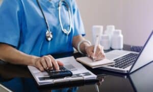 Medical professional calculating costs at a desk with a stethoscope around their neck. They are using a calculator, writing in a notebook, and working on a laptop, with medication bottles visible in the background.