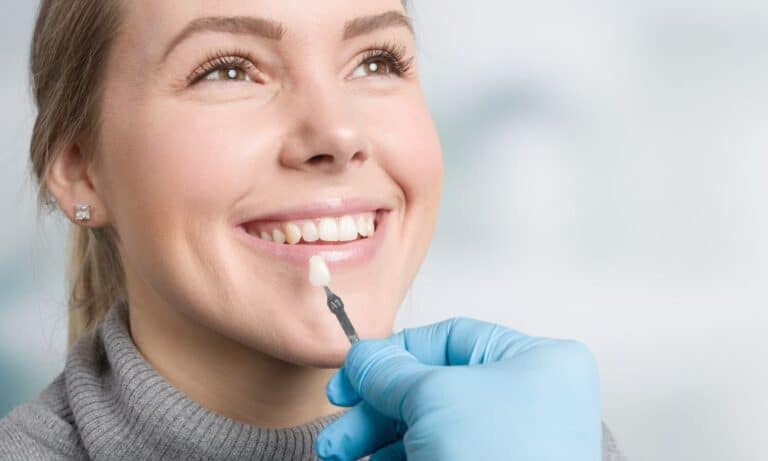 A woman with a bright smile is having a veneer shade matched to her teeth by a dentist wearing blue gloves. The background is blurred, focusing on the patient's happy expression and the dental procedure. The image highlights the process of getting veneers.