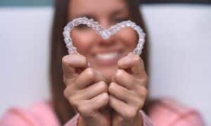 A woman holding Invisalign® aligners shaped into a heart, smiling in the background. The focus is on the clear aligners, illustrating their flexibility and design. The image represents the cost and value of Invisalign® treatment in the US.