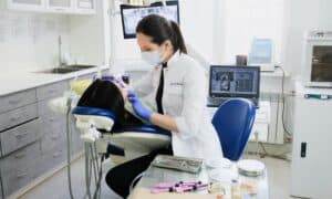 Dentist performing a dental procedure on a patient in a modern clinic. The dentist is wearing a mask and gloves, working in a well-equipped dental office with various tools and dental equipment. A computer monitor displays dental images in the background.