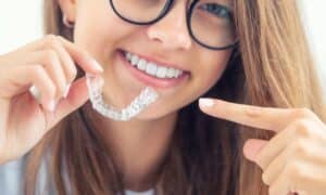 A young woman with glasses smiling and holding an Invisalign aligner, pointing to it with her other hand, showcasing the clear and discreet nature of the orthodontic treatment.