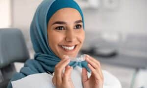 A smiling woman wearing a hijab holds an Invisalign aligner in a dental office, showcasing the effectiveness of Invisalign® for fixing crooked teeth.
