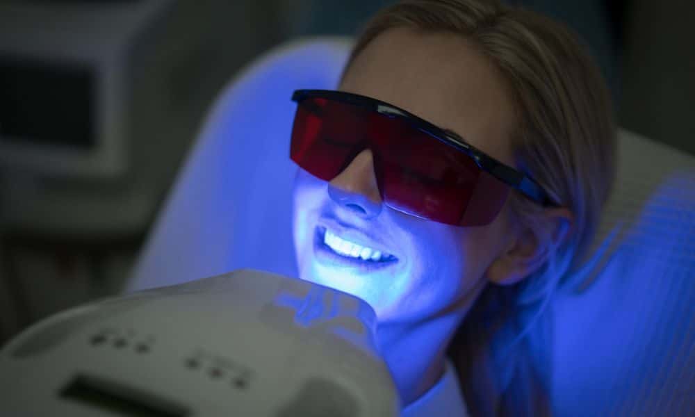 A woman undergoing professional teeth whitening treatment, wearing protective glasses, and smiling under a blue light. The image highlights the process and equipment used in professional teeth whitening, related to the cost of such treatments.