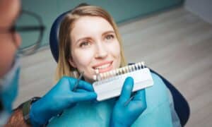 Dentist showing a patient a shade guide to match the color for veneers. The patient is smiling and sitting in a dental chair, while the dentist, wearing gloves and holding dental tools, helps her choose the right shade. The background is a dental clinic.
