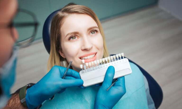 Dentist showing a patient a shade guide to match the color for veneers. The patient is smiling and sitting in a dental chair, while the dentist, wearing gloves and holding dental tools, helps her choose the right shade. The background is a dental clinic.