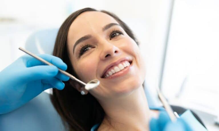 A woman with dark hair smiles while sitting in a dental chair during a check-up. A dentist wearing blue gloves holds a dental mirror near her mouth. The setting is bright and clean, emphasizing the patient's happy and confident expression.
