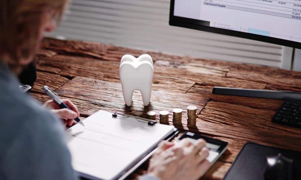 Person calculating costs at a desk with a model of a tooth and stacks of coins. The person is using a calculator and writing on a clipboard, with a computer screen displaying financial information in the background.