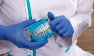 Dentist holding a model of teeth with braces, demonstrating orthodontic treatment. The dentist is wearing blue gloves and a white coat with a green trim.