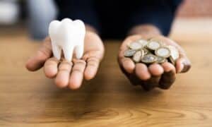 Person holding a model of a tooth in one hand and a handful of coins in the other, symbolizing the cost of dental implants. The background is a wooden table.
