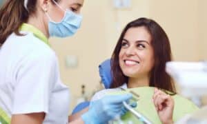 Happy woman sitting in a dental chair, smiling at her dentist. The dentist is holding dental tools and wearing a mask and gloves, preparing for the procedure.