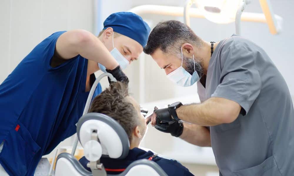 Dentists performing a dental implant procedure on a patient in a dental chair. Both dentists are wearing masks, gloves, and scrubs, focusing intently on the task.