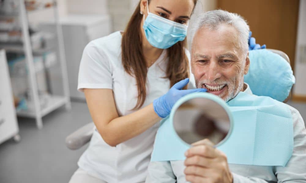 Elderly man smiling while a dentist shows him his new dental implants in a mirror. The dentist is wearing a mask and gloves, and the patient is sitting in a dental chair, looking happy with his results.