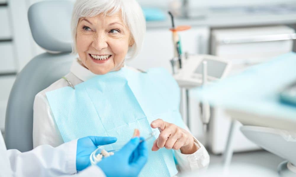Smiling elderly woman sitting in a dental chair, interacting with her dentist. The dentist is holding dental tools, and the patient appears pleased with the consultation.